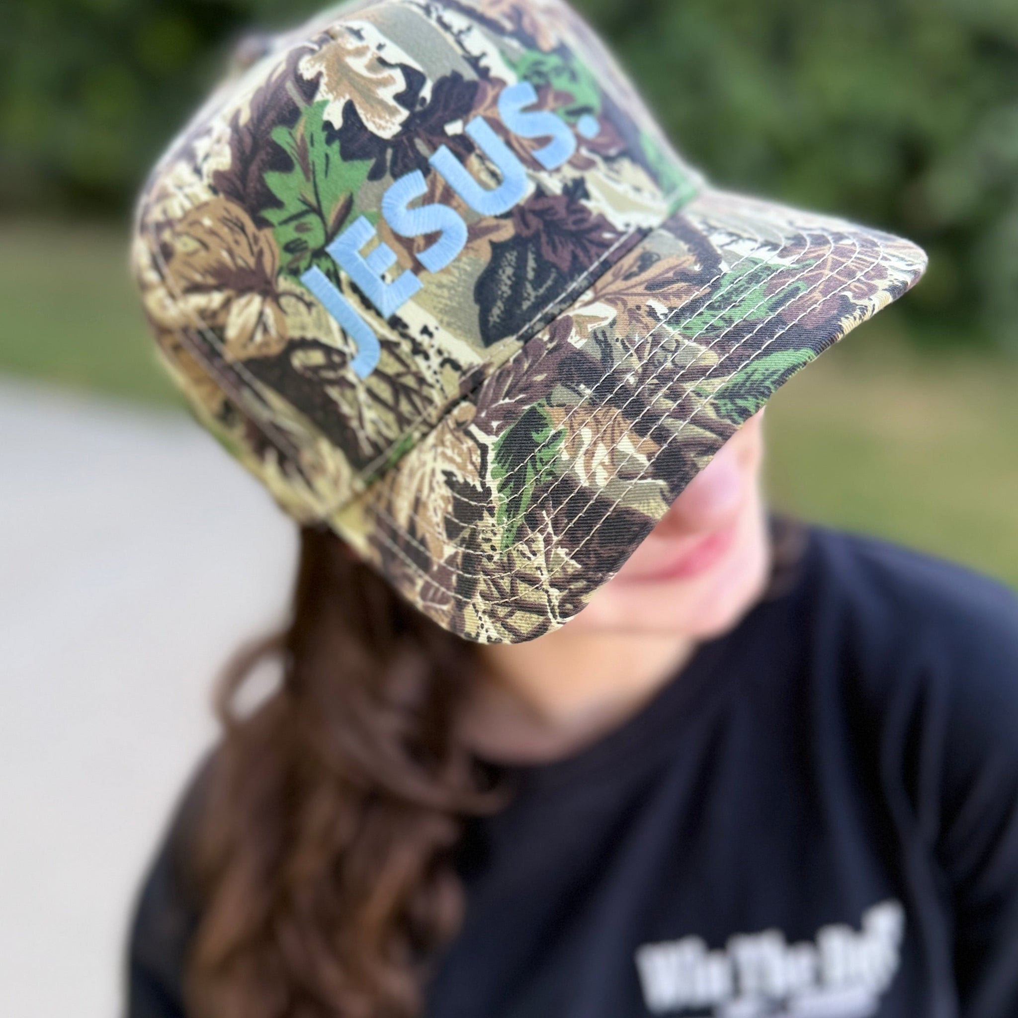 Person wearing a camouflage cap with 'JESUS' printed on it, outdoors.