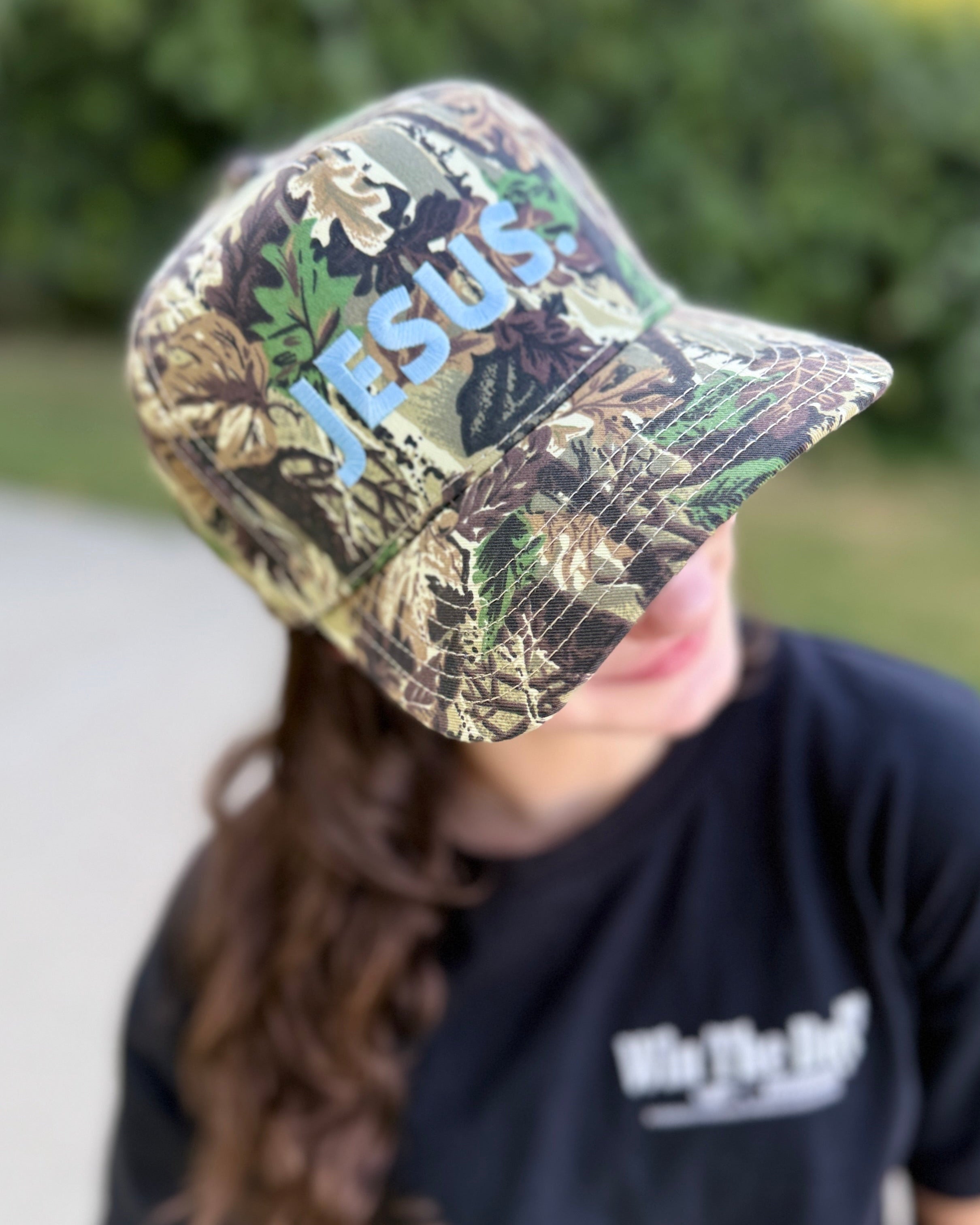 Person wearing a camouflage cap with 'JESUS' printed on it, outdoors.